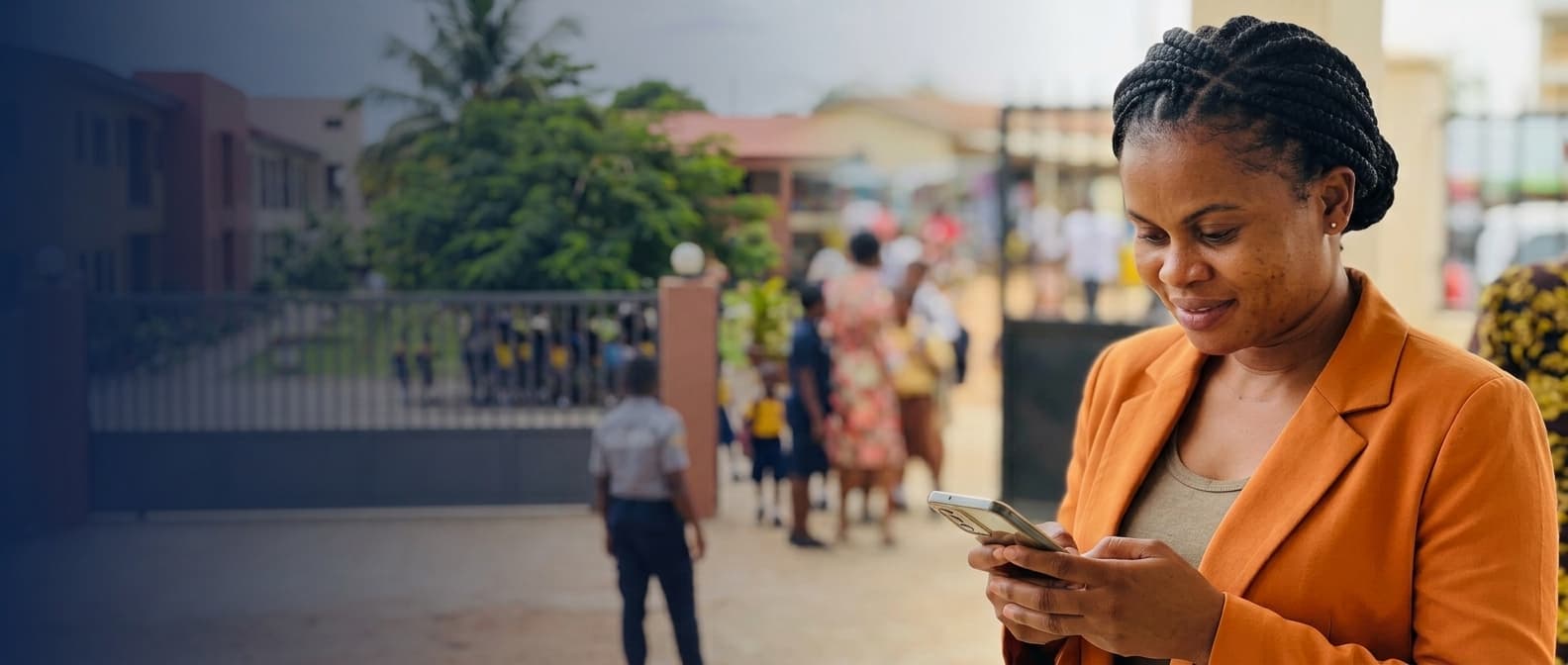 A school administrator checking her phone outside a school gate, with students and a security guard in the background